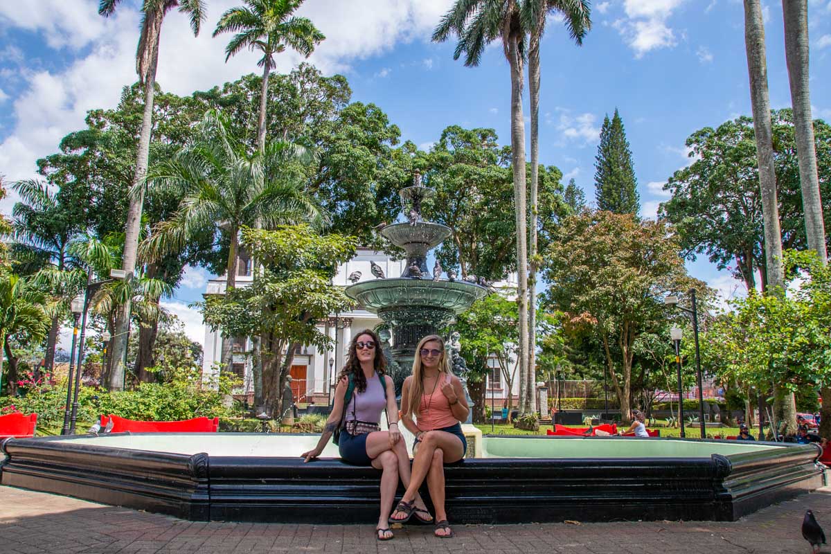 Two people sit in Alajuela Plaza at the fountain on a beautiful day