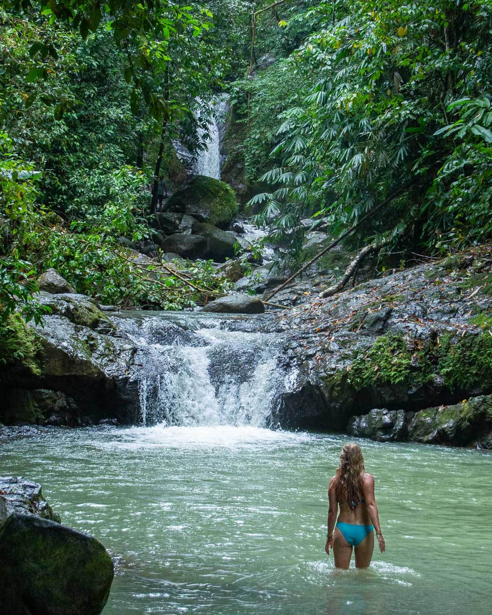 A lady stands in the water in the river below Uvita Waterfall with a view of the falls