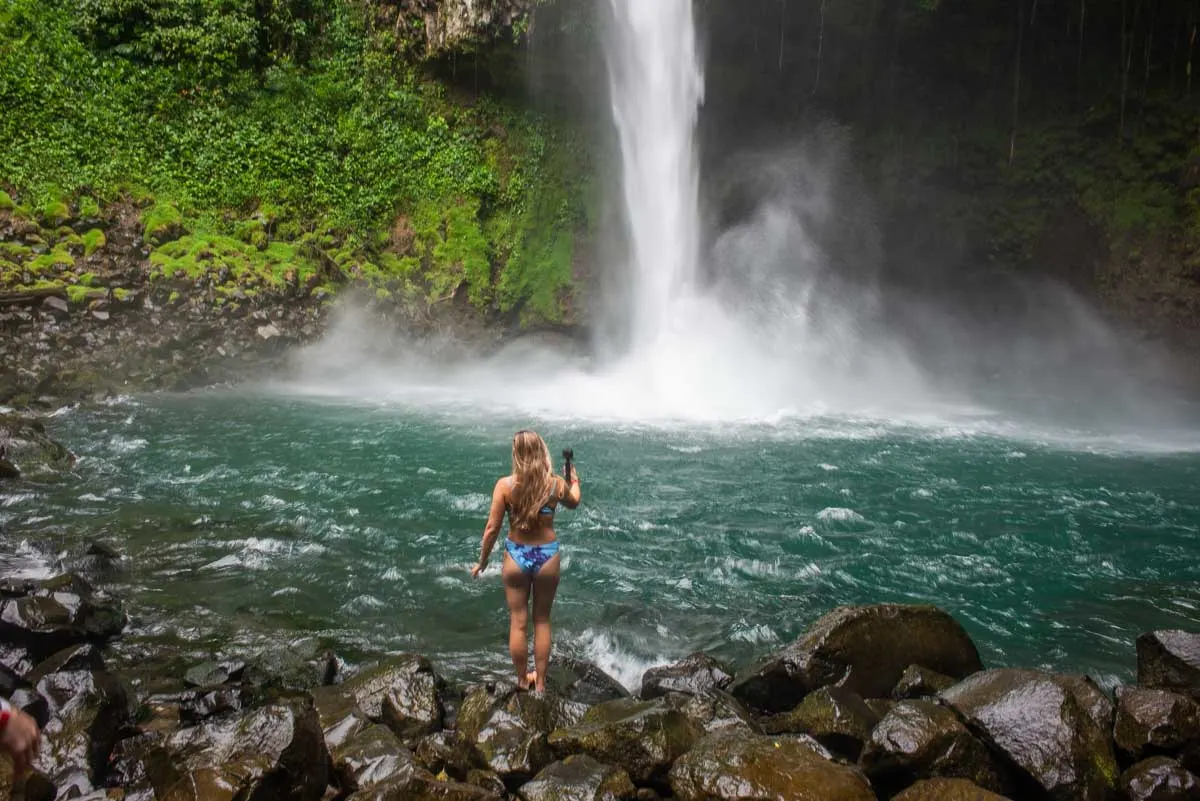 A lady walks towards the water with a goi pro at La Fortuna Waterfall