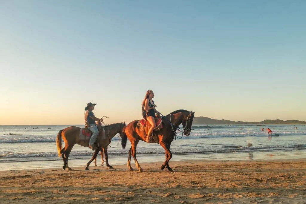 two people riding horses on a beach in Santa Teresa at sunset